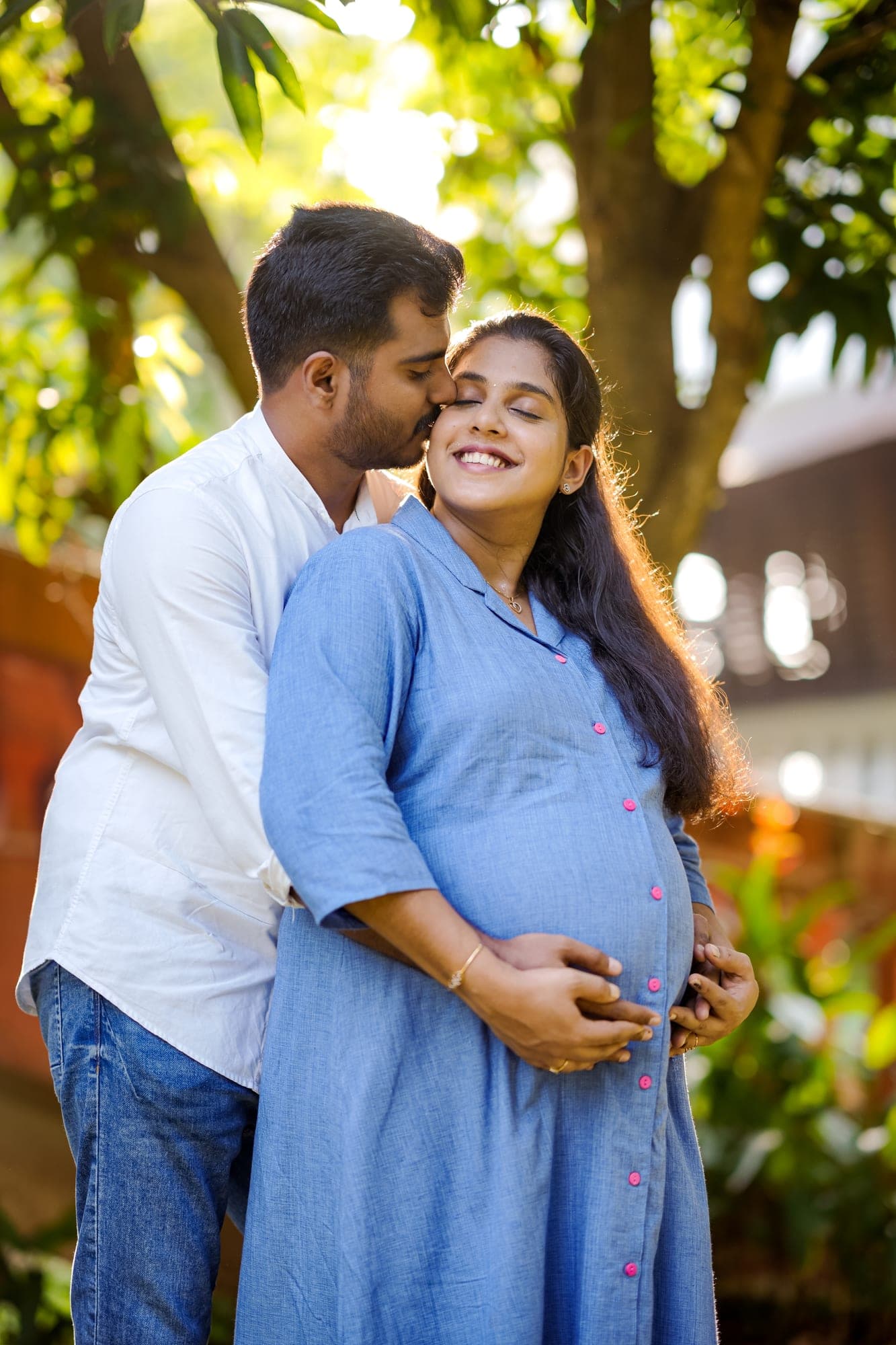 Husband kissing expecting wife's forehead under a tree