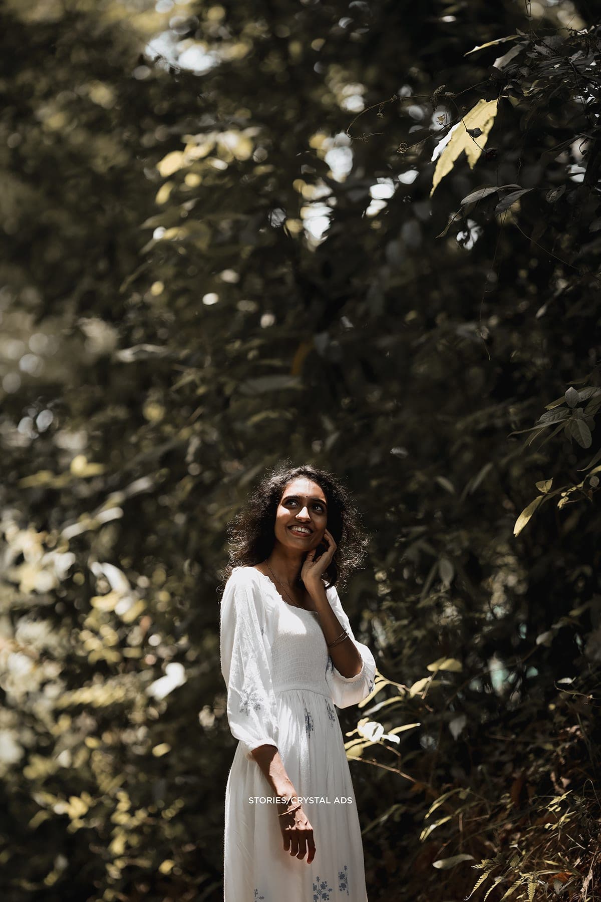 She stands among dark foliage in a white dress